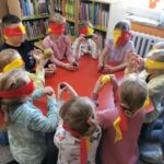 Spotkania dzieci z biblioteką - dzieci siedzą przy stole z przewiązanymi oczami i próbują odgadnąć jaki mają w ręku owoc bez używania wzroku