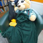 Śpiący na fotelu w bibliotece pluszowy misiu, przykryty zielonym kocem, obok niego leży miodek
