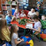 Dzieci w bibliotece siedzą przy stole, na stole jesienne warzywa, jedna z dziewczynek siedzących przy stole czyta pozostałym dzieciom z otwartej książki