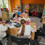 Troje dzieci przy czerwonym stoliku w bibliotece wykonuje swoje prace plastyczne - papierowe pojemniki w kształcie wielkanocnego zajączka