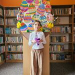 Dziewczynka na tle półek w bibliotece prezentuje wykonaną pracę plastyczną - papierowy pojemnik w kształcie wielkanocnego zajączka