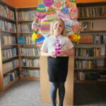 Dziewczynka na tle półek w bibliotece prezentuje wykonaną pracę plastyczną - papierowy pojemnik w kształcie wielkanocnego zajączka