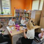 Dzieci przy stoliku w bibliotece wykonuje swoje prace plastyczne - papierowe pojemniki w kształcie wielkanocnego zajączka
