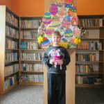 Dziewczynka na tle półek w bibliotece prezentuje wykonaną pracę plastyczną - papierowy pojemnik w kształcie wielkanocnego zajączka