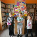 Troje dzieci na tle półek w bibliotece prezentuje wykonaną pracę plastyczną - papierowy pojemnik w kształcie wielkanocnego zajączka