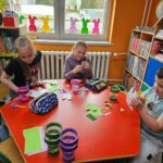 Troje dzieci przy czerwonym stoliku w bibliotece wykonuje swoje prace plastyczne - papierowe pojemniki w kształcie wielkanocnego zajączka