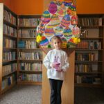 Dziewczynka na tle półek w bibliotece prezentuje wykonaną pracę plastyczną - papierowy pojemnik w kształcie wielkanocnego zajączka