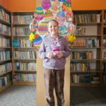 Dziewczynka na tle półek w bibliotece prezentuje wykonaną pracę plastyczną - papierowy pojemnik w kształcie wielkanocnego zajączka