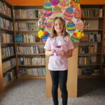 Dziewczynka na tle półek w bibliotece prezentuje wykonaną pracę plastyczną - papierowy pojemnik w kształcie wielkanocnego zajączka