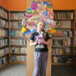 Dziewczynka na tle półek w bibliotece prezentuje wykonaną pracę plastyczną - papierowy pojemnik w kształcie wielkanocnego zajączka