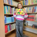 Dziewczynka prezentuje wykonaną przez siebie pracę plastyczną stojąc na tle bibliotecznych półek