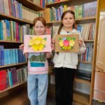 Dwie dziewczynki prezentują wykonane przez siebie prace plastyczne stojąc na tle bibliotecznych półek
