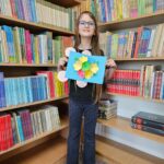 Dziewczynka prezentuje wykonaną przez siebie pracę plastyczną stojąc na tle bibliotecznych półek