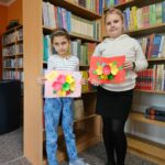 Dwie dziewczynki prezentują wykonane przez siebie prace plastyczne stojąc na tle bibliotecznych półek