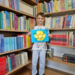 Dziewczynka prezentuje wykonaną przez siebie pracę plastyczną stojąc na tle bibliotecznych półek