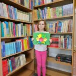 Dziewczynka prezentuje wykonaną przez siebie pracę plastyczną stojąc na tle bibliotecznych półek