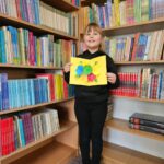Dziewczynka prezentuje wykonaną przez siebie pracę plastyczną stojąc na tle bibliotecznych półek