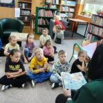 Dzieci siedzą na podłodze biblioteki i z uwagą słuchają czytanego opowiadania