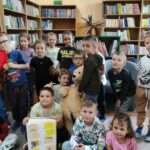 Pamiątkowe dzieci siedzące na podłodze biblioteki a pośrodku nich, trzymana przez jedno z dzieci wykonana praca - przedstawiająca grubą książkę z naklejonymi wcześniej wykonanymi "dymkami"