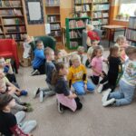 Dzieci siedzą i klęczą na podłodze biblioteki i z uwagą słuchają czytanego opowiadania