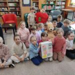 Pamiątkowe dzieci siedzące na podłodze biblioteki a pośrodku nich., trzymana przez troje dzieci wykonana praca - przedstawiająca grubą książkę z naklejonymi "dymkami"