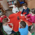 Sześcioro dzieci siedzą przy sześciokątnym, czerwonym stole, na środku którego znajduje się biurowy pojemnik z kredkami