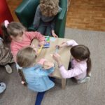 Czworo dzieci klęcząc przy niskim, kwadratowym stole wykonuje prace plastyczne - na karteczkach w kształcie komiksowych "dymków" rysuje swoje wspomnienia z przedszkola