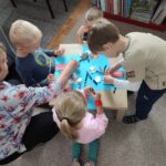 Dzieci wraz z panią przy stole w bibliotece wykonują swoją pracę plastyczną - bocianie gniazda, wykorzystując kolorowe kartki, płatki kosmetyczne i klej