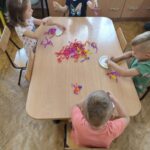 Dzieci przy stolikach wykonują pracę plastyczną - z tekturowego talerzyka przeciętego na pół i doklejonych do niego nóżek z kolorowej bibuły - tworzą "ośmiornice"