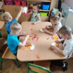 Dzieci przy stolikach wykonują pracę plastyczną - z tekturowego talerzyka przeciętego na pół i doklejonych do niego nóżek z kolorowej bibuły - tworzą "ośmiornice"