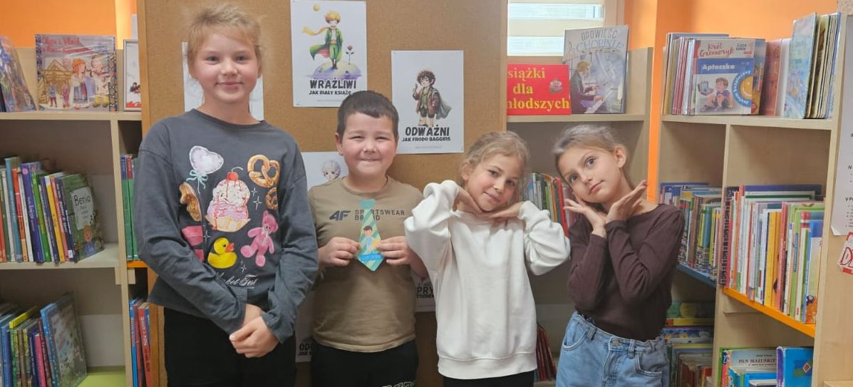 Grupa dzieci w sali biblioteki stoi na tle półek z książkami