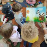 Grupa dzieci z uwagą słucha wierszyka czytanego przez Panią w bibliotece.