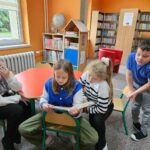 Dziewczynka czyta opowiadanie w sali biblioteki, a dzieci obok niej z uwagą słuchają