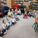 Dzieci siedzą na podłodze po turecku w sali bibliotecznej i z uwagą słuchają opowiadania