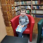 Przedszkolak siedzi w czerwonym fotelu w sali biblioteki a w ręku trzyma książkę