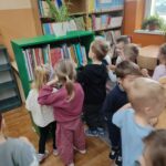 Grupa przedszkolaków stoi przed półką z książkami, przygotowanymi specjalnie dla nich przez Panie z biblioteki