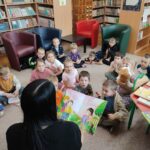 Dzieci siedzą na podłodze sali biblioteki i aktywnie uczestniczą w zajęciach