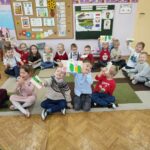 Zdjęcie grupowe przedszkolaków siedzących na podłodze i prezentujących wykonane przez sieie prace plastyczne