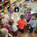 Dzieci stoją i wspólnie trzymają duży arkusz szarego papieru, na którym na narysowanych czarnym flamastrem torach przeklejona jest lokomotywa i 14 wagoników - każdy zrobiony przez uczestnika spotkania