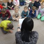 Przedszkolaki siedzą na podłodze i z uwagą słuchają opowiadania czytanego przez nauczyciela