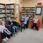 Przedszkolaki siedzą na fotelach w bibliotece i z uwagą słuchają opowiadania czytanego przez nauczyciela
