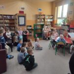Dzieci siedzą na ziemi i z uwagą słuchają czytanego przez panią opowiadania