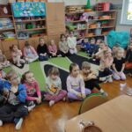 Dzieci siedzą na ziemi i z uwagą słuchają czytanego przez panią opowiadania