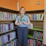 Dziewczynka stoi na tle bibliotecznych regałów z książkami i prezentuje wykonaną przez siebie pracę