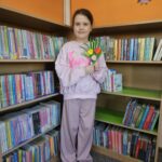 Dziewczynka stoi na tle bibliotecznych regałów z książkami i prezentuje wykonaną przez siebie pracę
