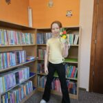 Dziewczynka stoi na tle bibliotecznych regałów z książkami i prezentuje wykonaną przez siebie pracę