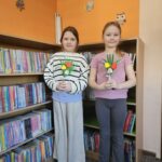 Dziewczynki stoją na tle bibliotecznych regałów z książkami i prezentują wykonaną przez siebie pracę