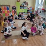 Dzieci siedzą na ziemi i z uwagą słuchają czytanego przez panią opowiadania