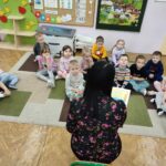 Dzieci siedzą na ziemi i z uwagą słuchają czytanego przez panią opowiadania