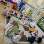 Dzieci siedzą na ziemi i z uwagą słuchają czytanego przez panią opowiadania